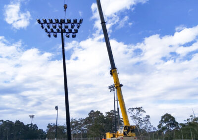 GM Poles 25m black powdercoated lighting tower in the process of being installed. The pole is being pulled up off the ground by a large crane.