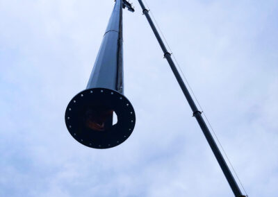 GM Poles Black powdercoated 25m lighting tower is suspended in the air by a large crane boom. The photo shows the pole from below with a blue sky as the background